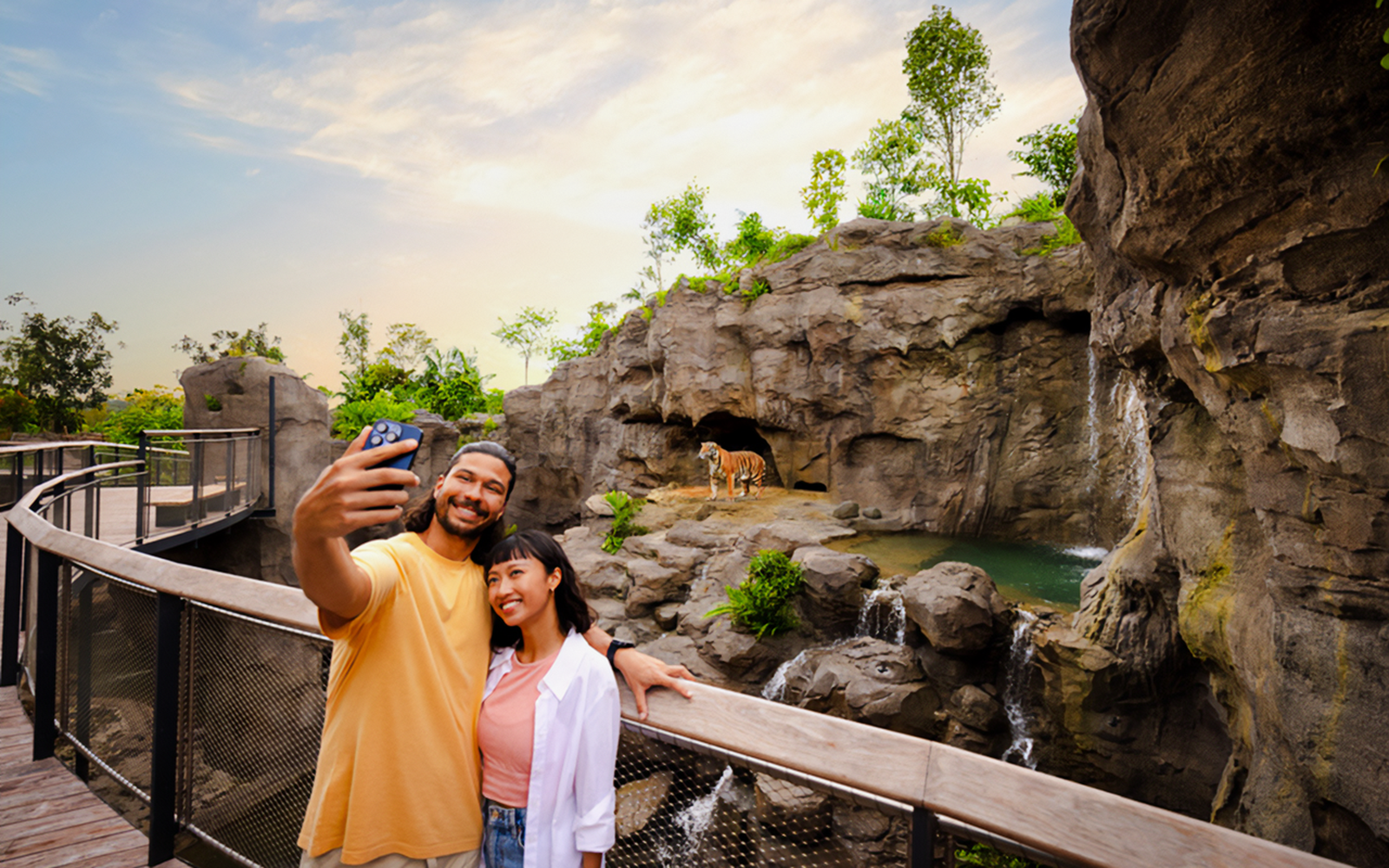 Couple taking selfie near rock cascade with tiger in background at zoo.
