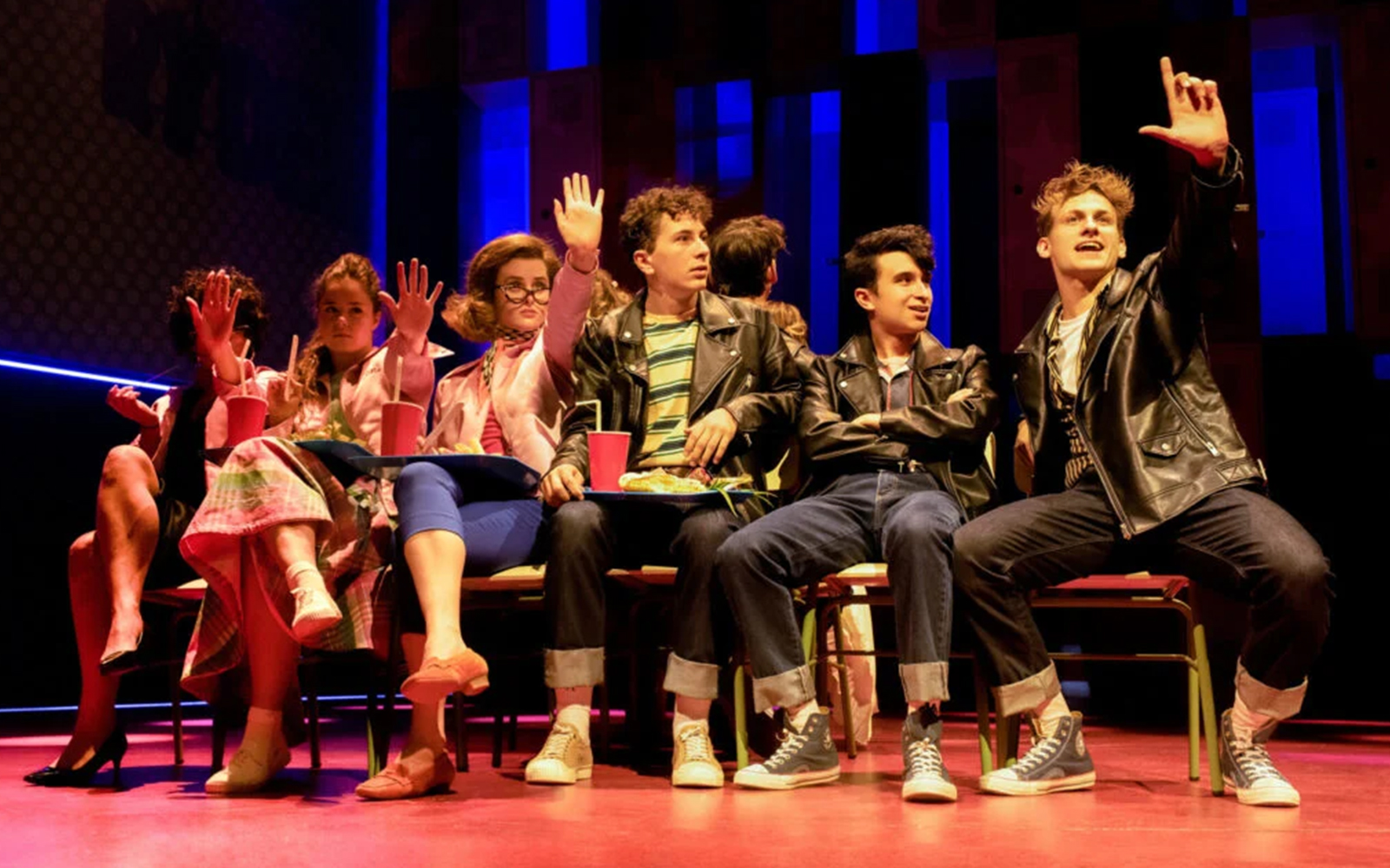 Cast members in leather jackets and pink outfits performing a scene from Grease on stage.