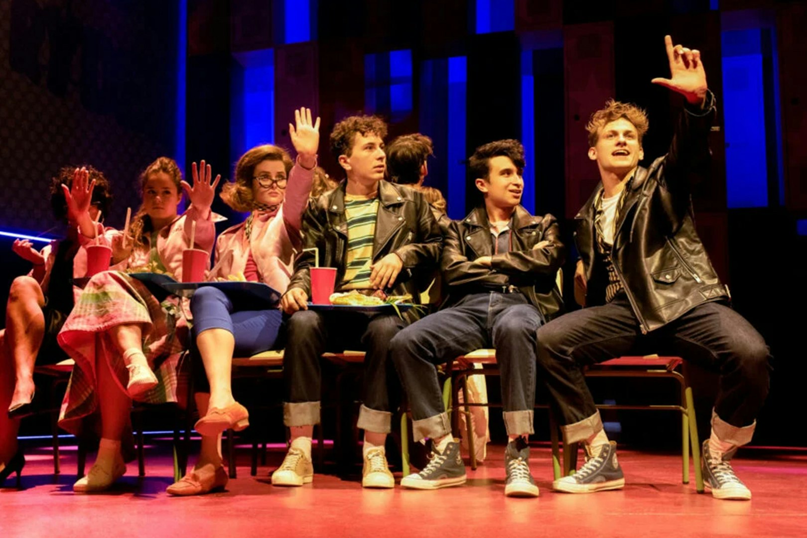Cast members in leather jackets and pink outfits performing a scene from Grease on stage.
