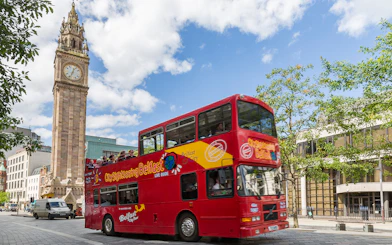 Red double-decker bus on Belfast Hop-On Hop-Off Tour passing Albert Memorial Clock.