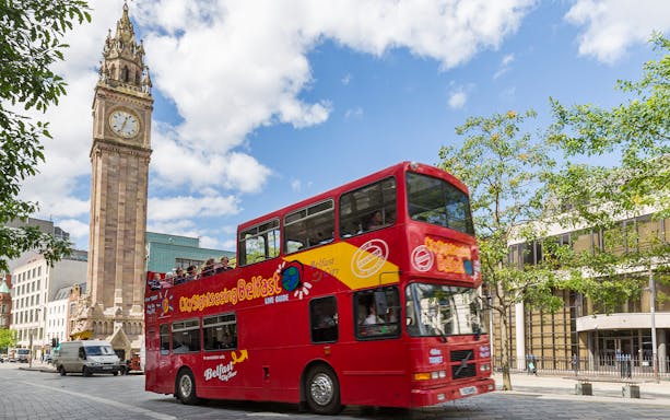Red double-decker bus on Belfast Hop-On Hop-Off Tour passing Albert Memorial Clock.