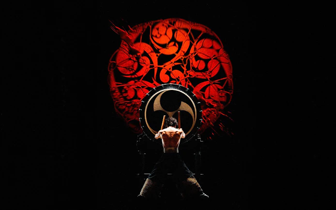 Performer playing taiko drum at DRUM TAO Theater Kyoto with red backdrop.
