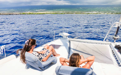 Two women relaxing on a catamaran cruise with ocean and Kona, Hawaii coastline in view.