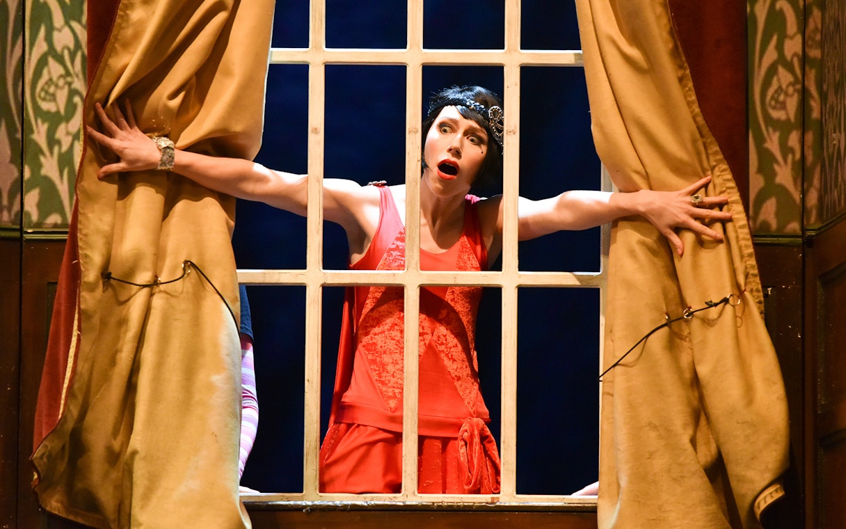 Performer in red dress on stage during "The Play That Goes Wrong" scene.