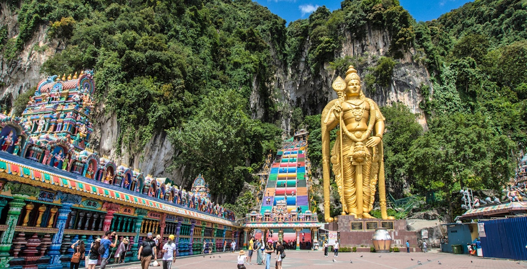 Golden statue and colorful temple at Batu Caves, Malaysia, with tourists exploring the site.