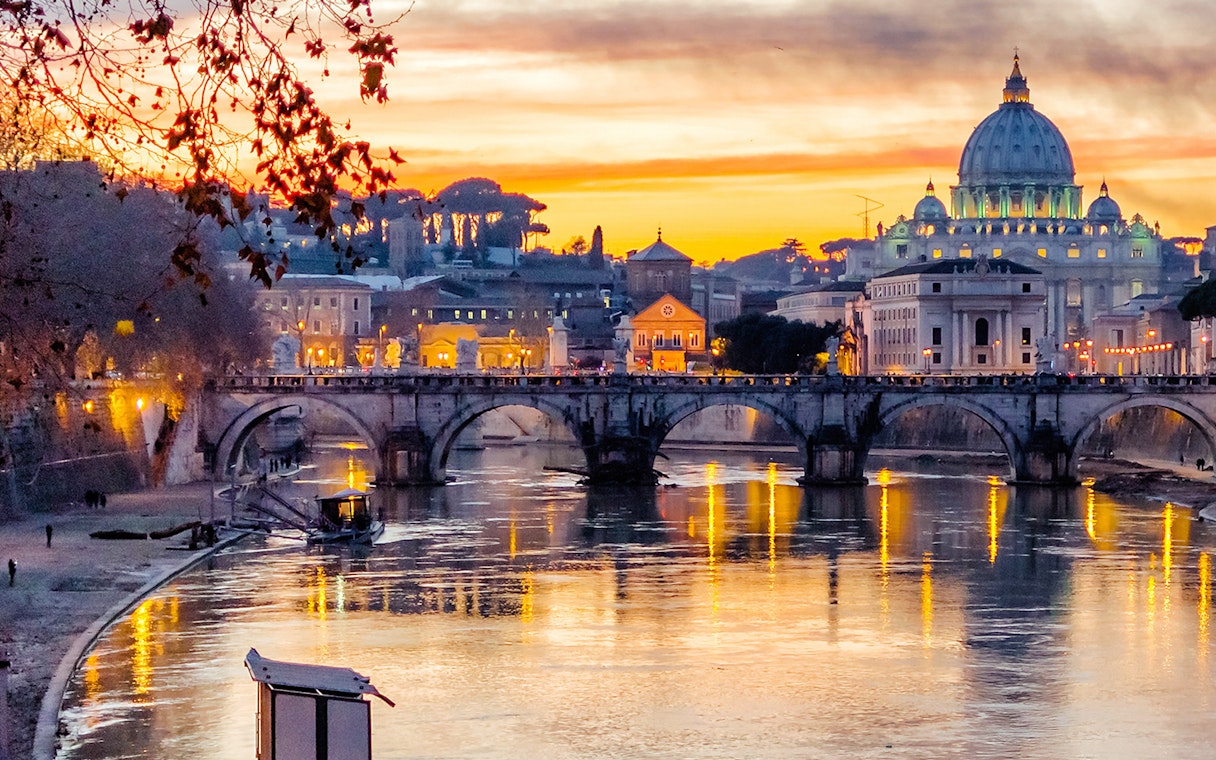 Vatican City skyline and St. Peter's Basilica at sunset, viewed from the Tiber River in Rome.