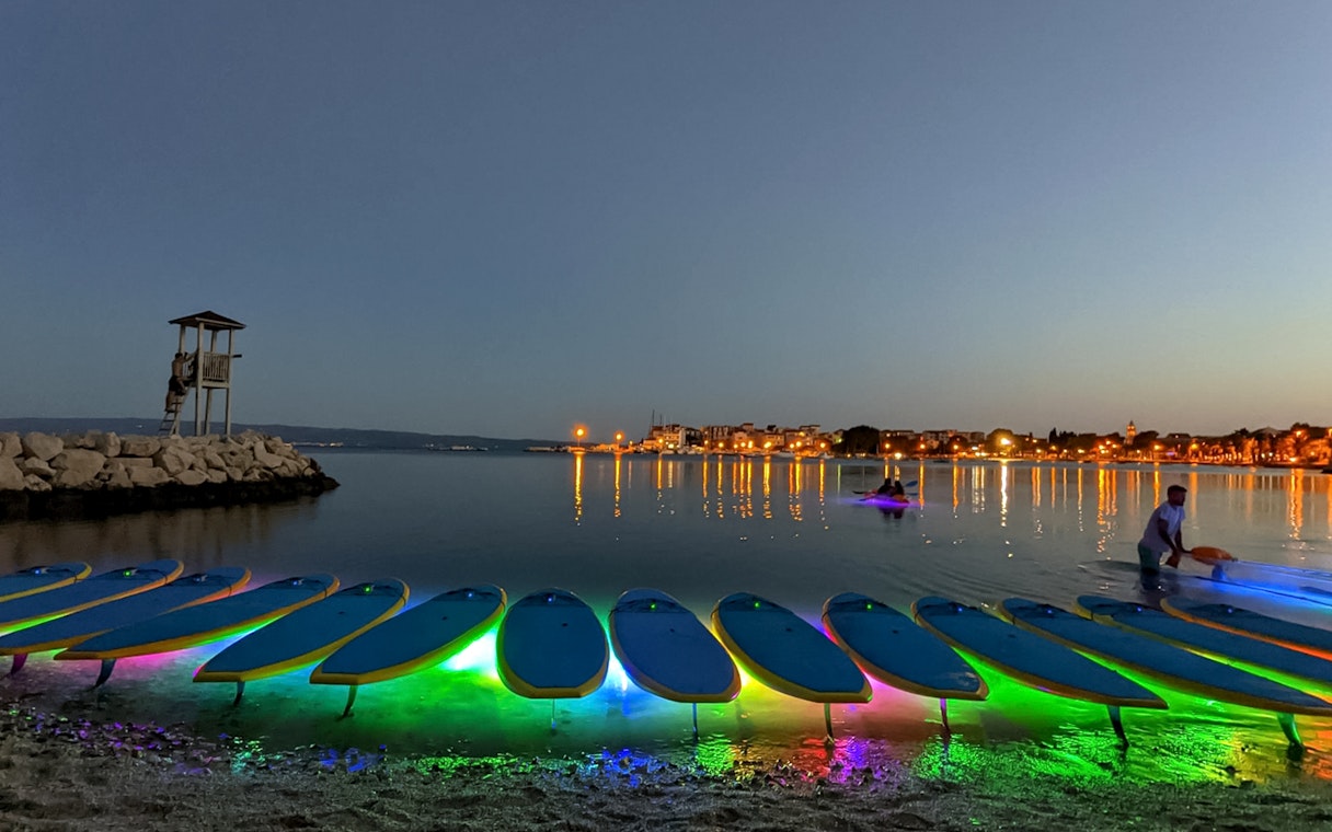 SUP boards with colorful lights on the shore at dusk during a Glow Tour in Split.