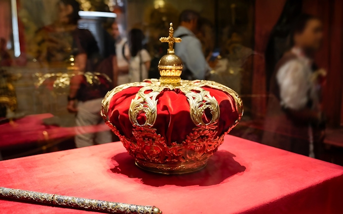 Royal crown on display at the Royal Palace of Madrid with tour group in background.