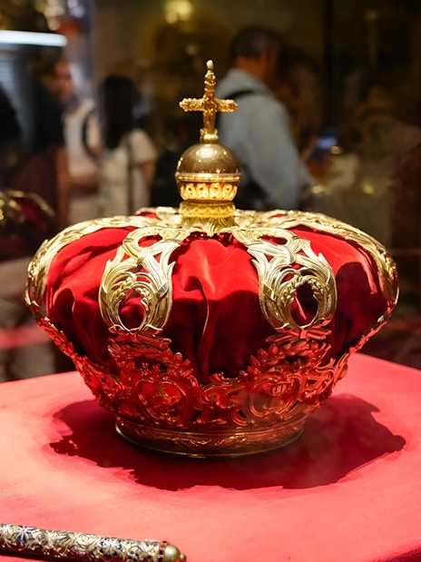 Royal crown on display at the Royal Palace of Madrid with tour group in background.