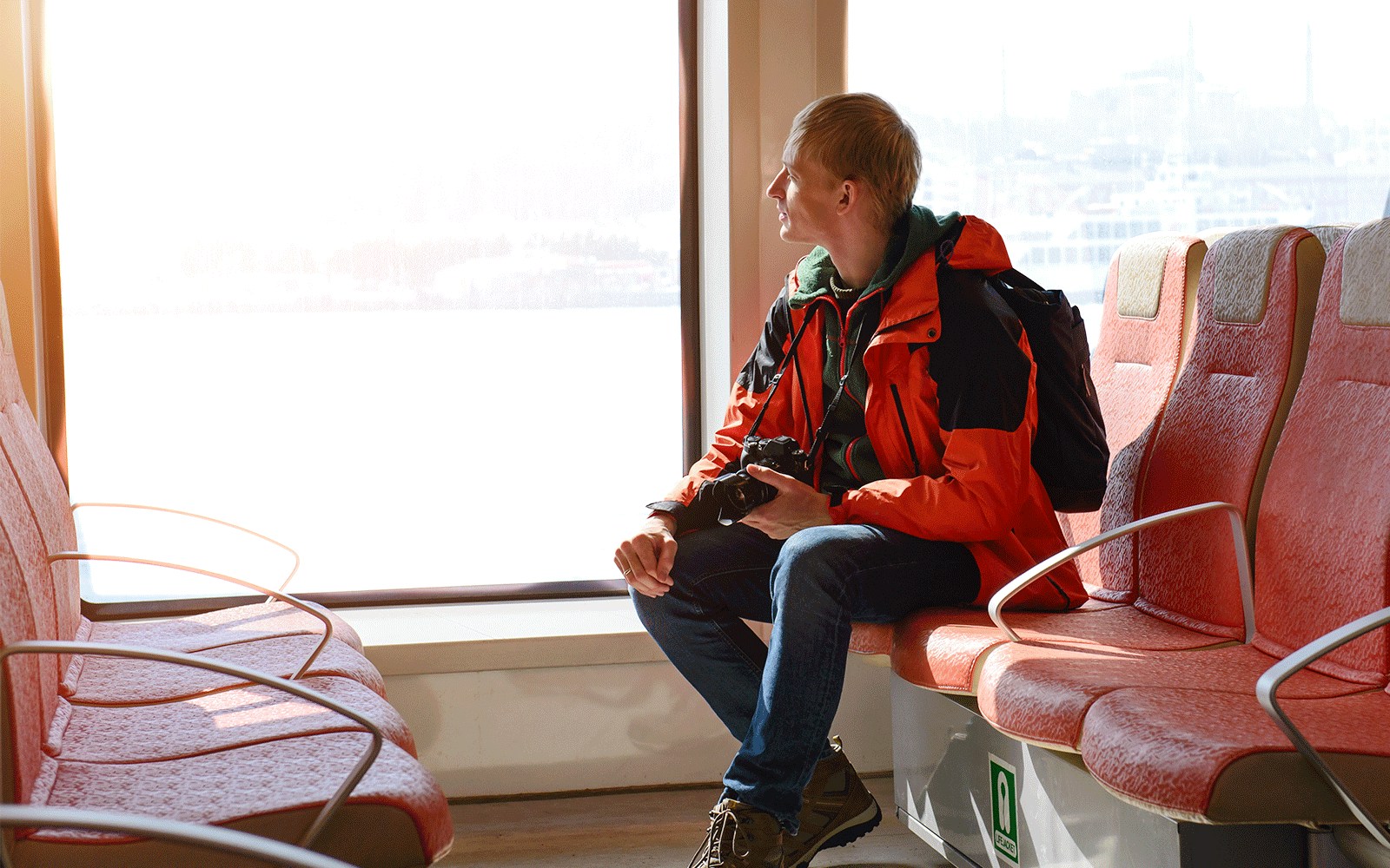 Passenger with camera seated comfortably on ferry, gazing out window.