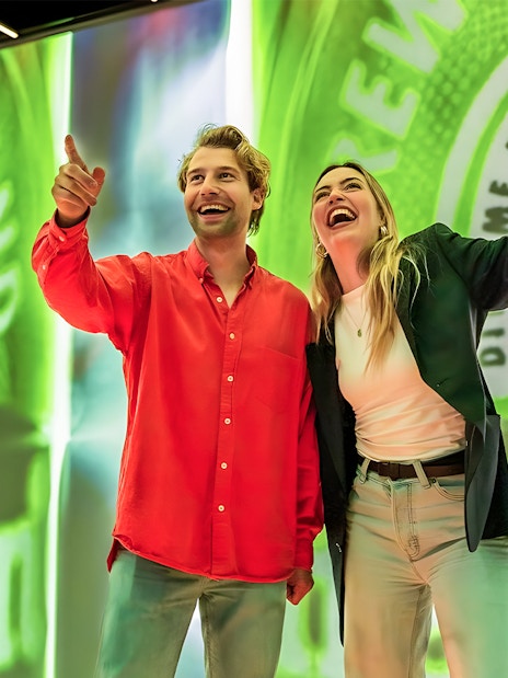 Tourists smiling and pointing at Heineken Experience in Amsterdam.