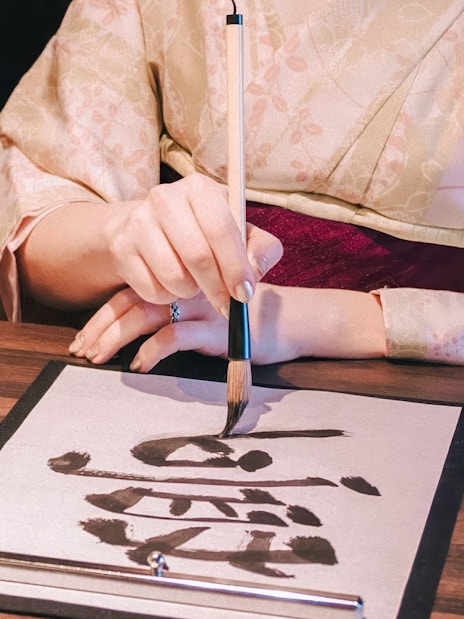 Kimono-clad person practicing Japanese calligraphy at a tea ceremony.