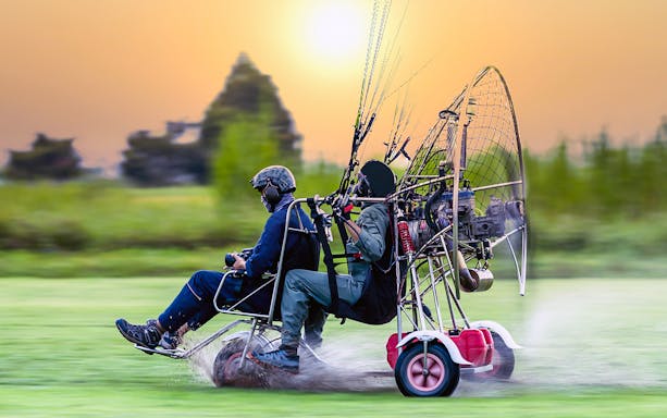 Tandem paramotor flight over grassy field at sunset.