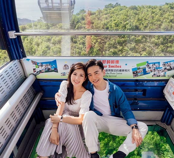 Couple enjoying view from crystal cabin cable car with glass floor over lush forest.