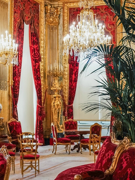 Palace of Versailles room with ornate chandeliers, red velvet chairs, and gold decor.