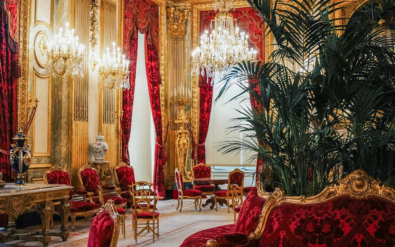 Palace of Versailles room with ornate chandeliers, red velvet chairs, and gold decor.