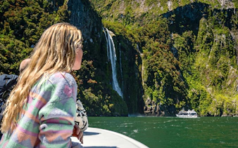 Tourist on boat viewing waterfall at Milford Sound, New Zealand, with lush cliffs and cruise ship in background.