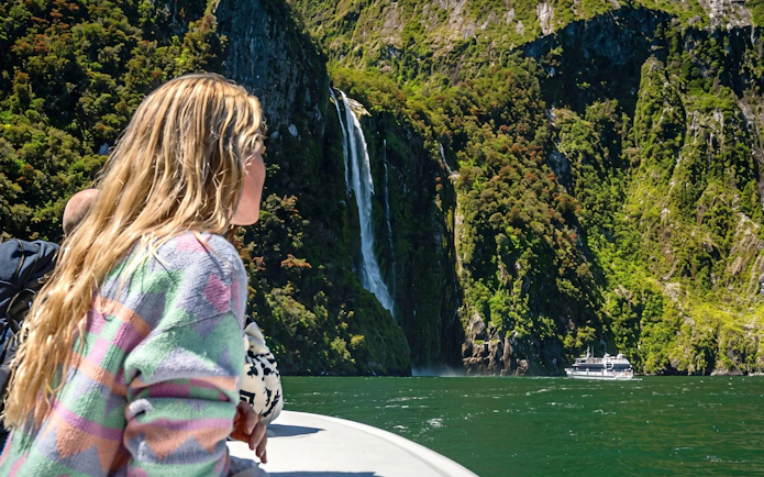 Tourist on boat viewing waterfall at Milford Sound, New Zealand, with lush cliffs and cruise ship in background.