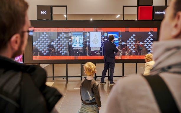 Interactive exhibit at Museum of Ethnography, Budapest, with visitors observing a digital timeline display.