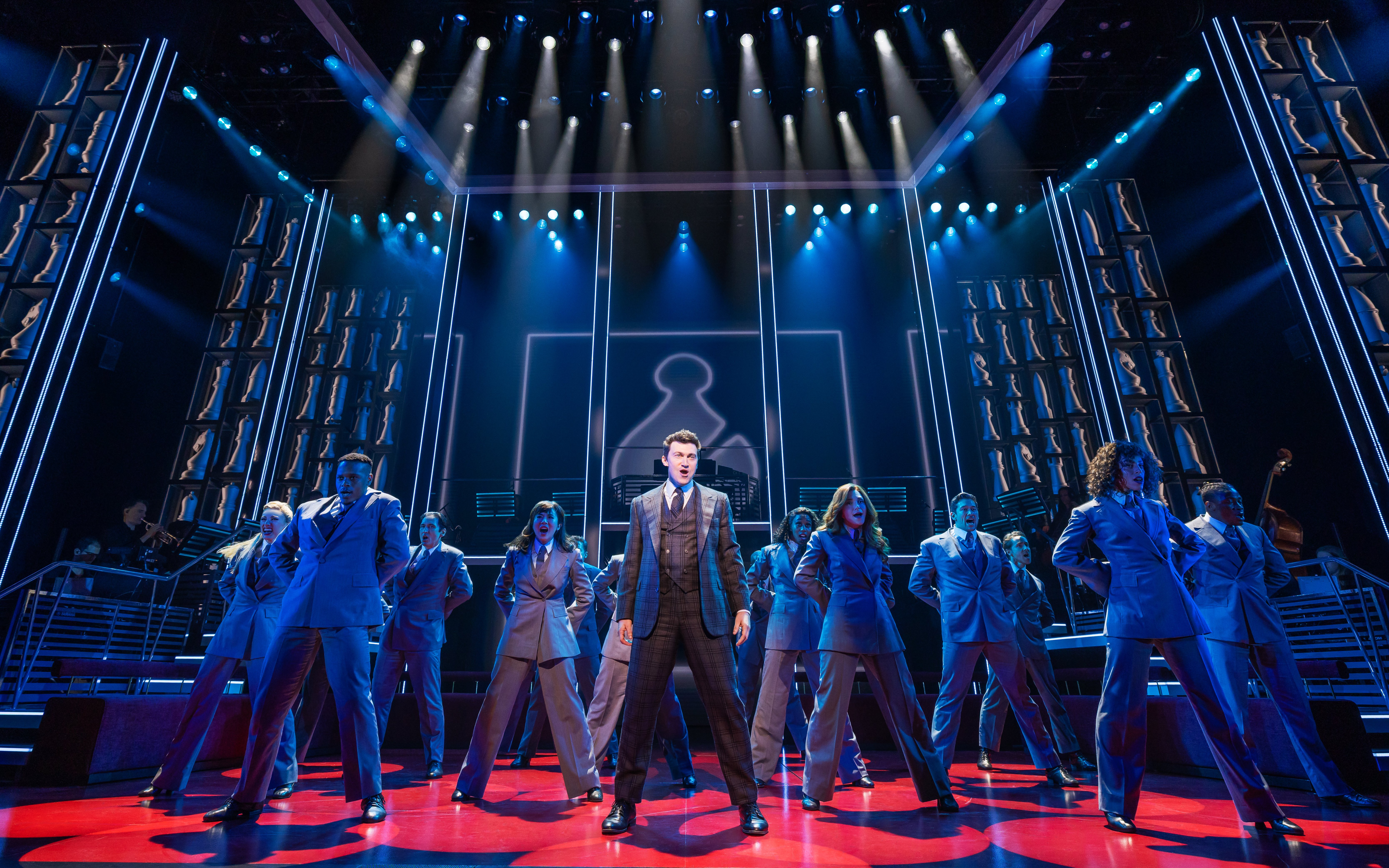 Stage performance of Chess Broadway show with cast in suits under blue lighting.