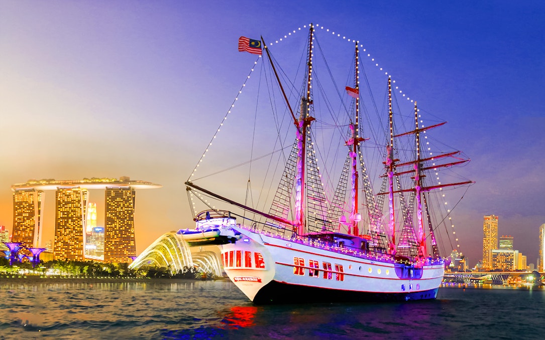 Sailing ship Royal Albatross illuminated at night with Singapore skyline in the background.