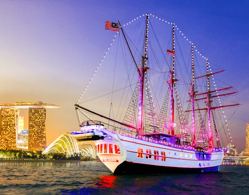 Sailing ship Royal Albatross illuminated at night with Singapore skyline in the background.