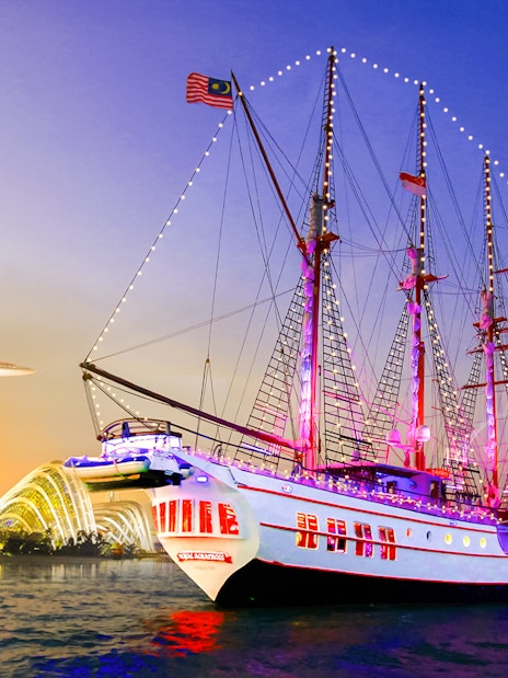 Sailing ship Royal Albatross illuminated at night with Singapore skyline in the background.