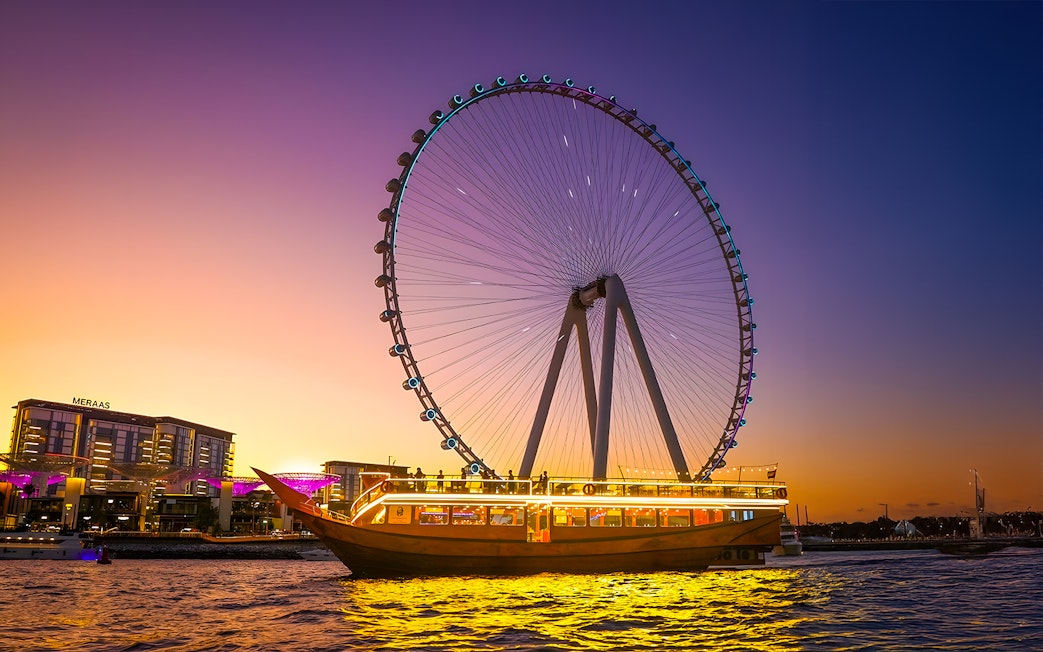 Dhow cruise with lights on Dubai Marina at sunset, near Ferris wheel.