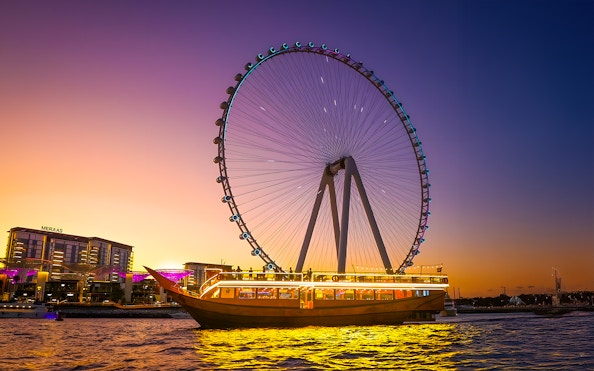 Dhow cruise with lights on Dubai Marina at sunset, near Ferris wheel.