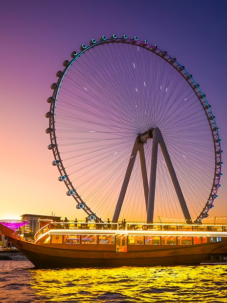 Dhow cruise with lights on Dubai Marina at sunset, near Ferris wheel.