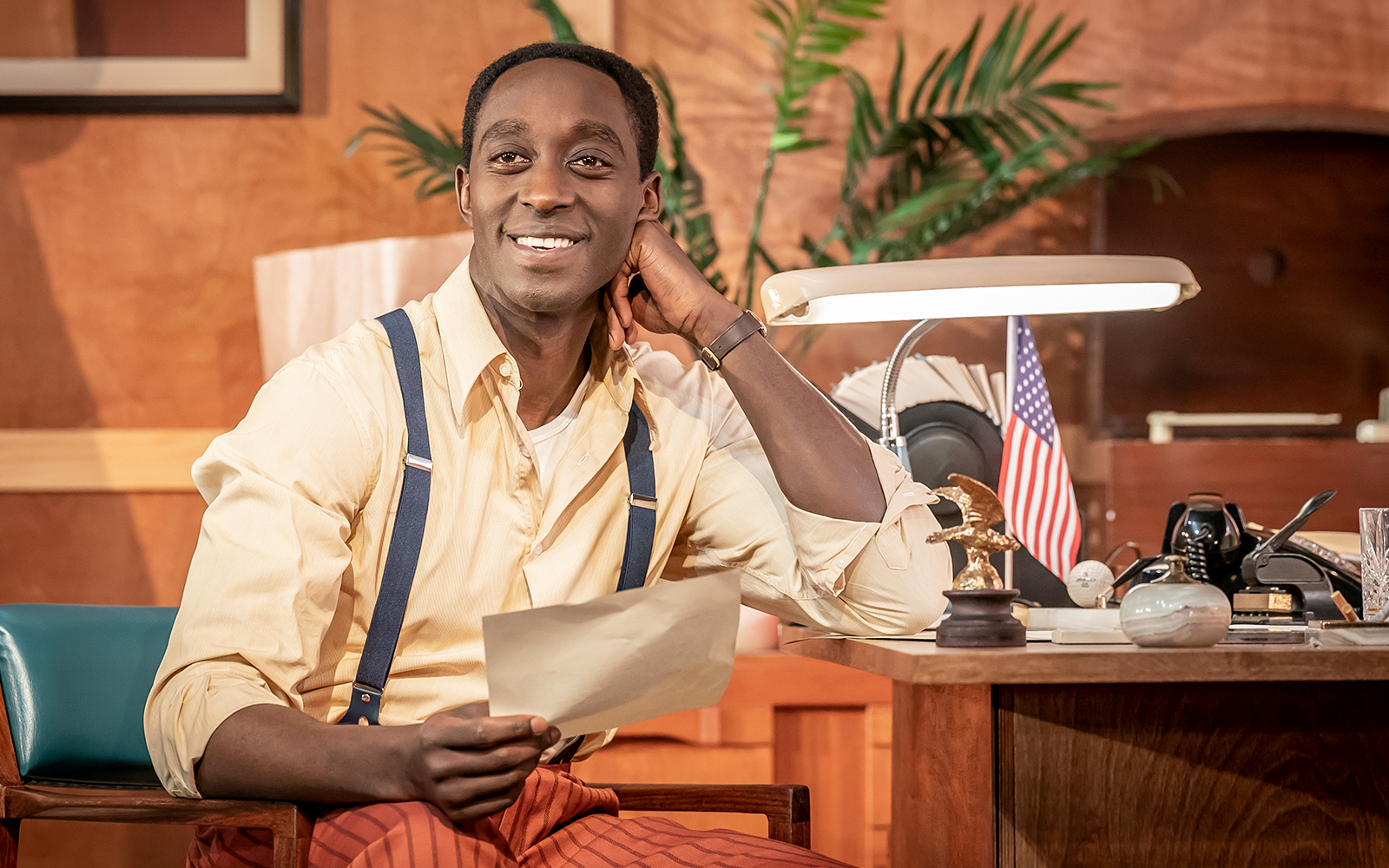 Man smiling at desk with vintage decor, holding paper, American flag in background.