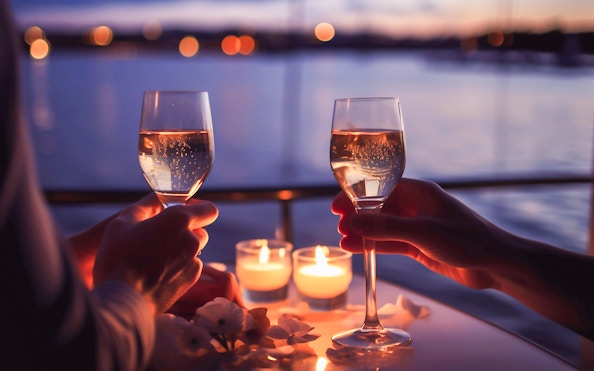 Two people toasting with wine glasses on a candlelit cruise at sunset.