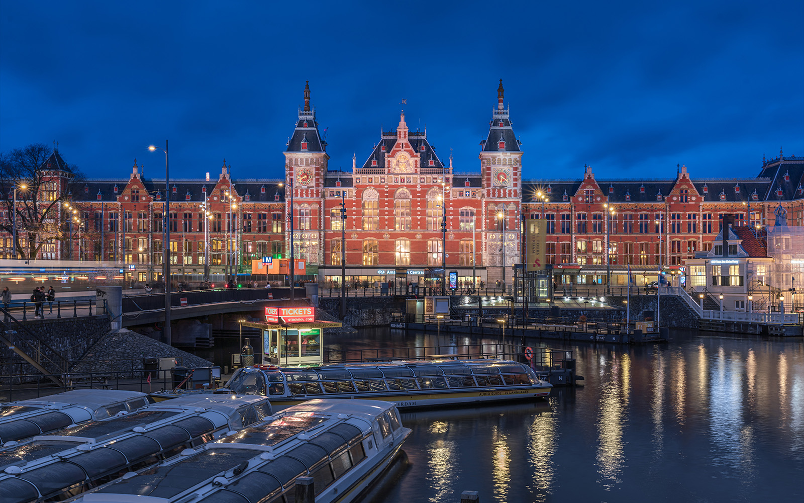 Amsterdam Centraal station illuminated at night with canal boats in the foreground.