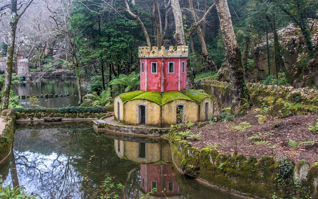 Small red and yellow tower by a pond in Pena Park, Sintra, surrounded by lush greenery.