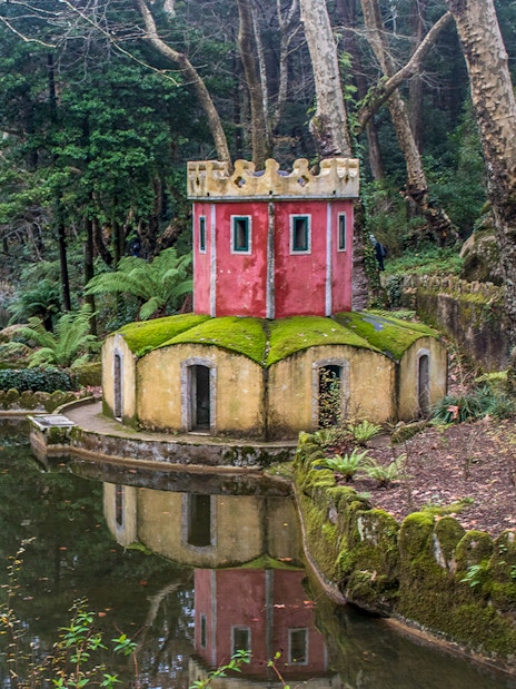 Small red and yellow tower by a pond in Pena Park, Sintra, surrounded by lush greenery.
