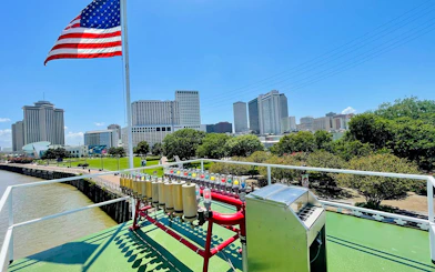 Riverboat deck with calliope and American flag, New Orleans skyline in background.