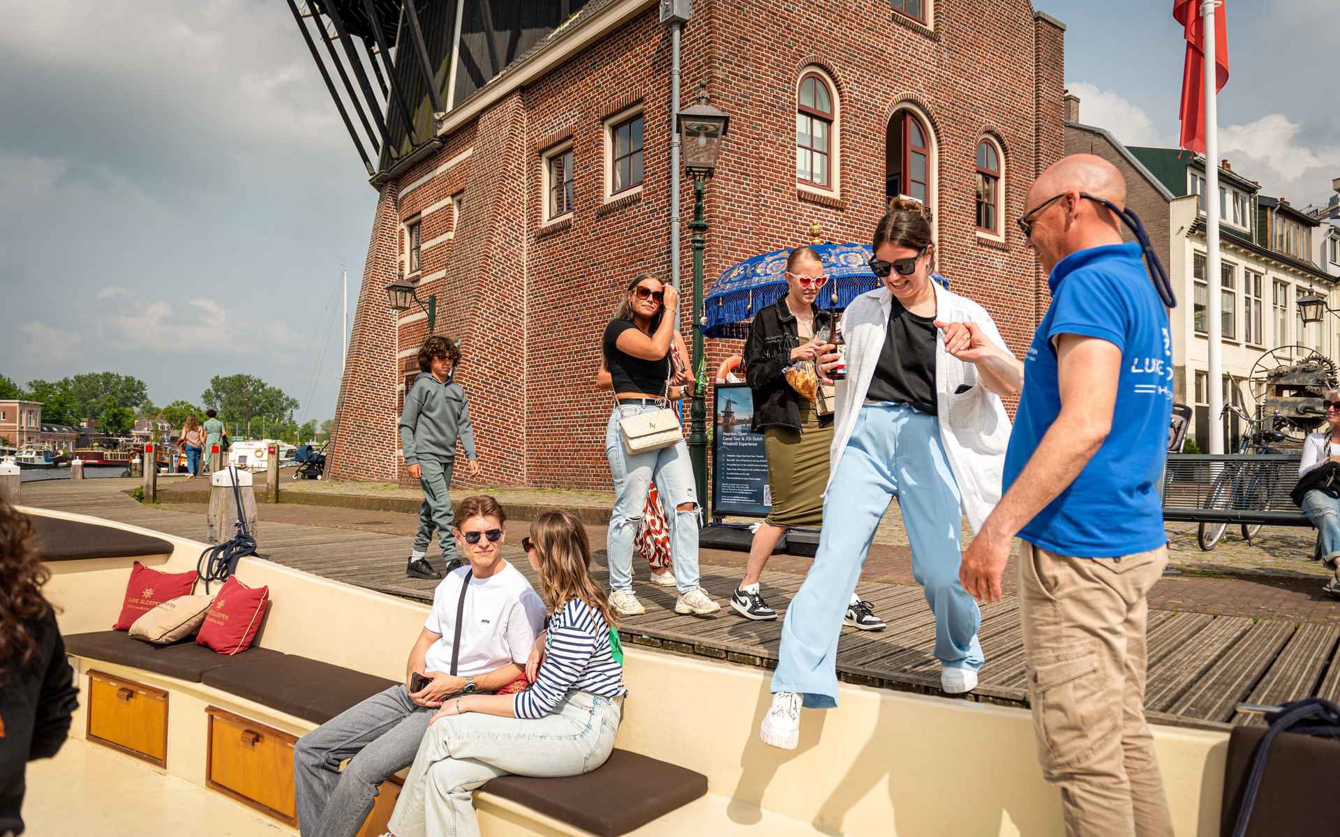 People boarding an open boat for a Harlem canal cruise near a historic brick building.