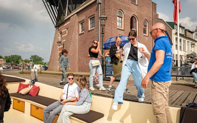 People boarding an open boat for a Harlem canal cruise near a historic brick building.