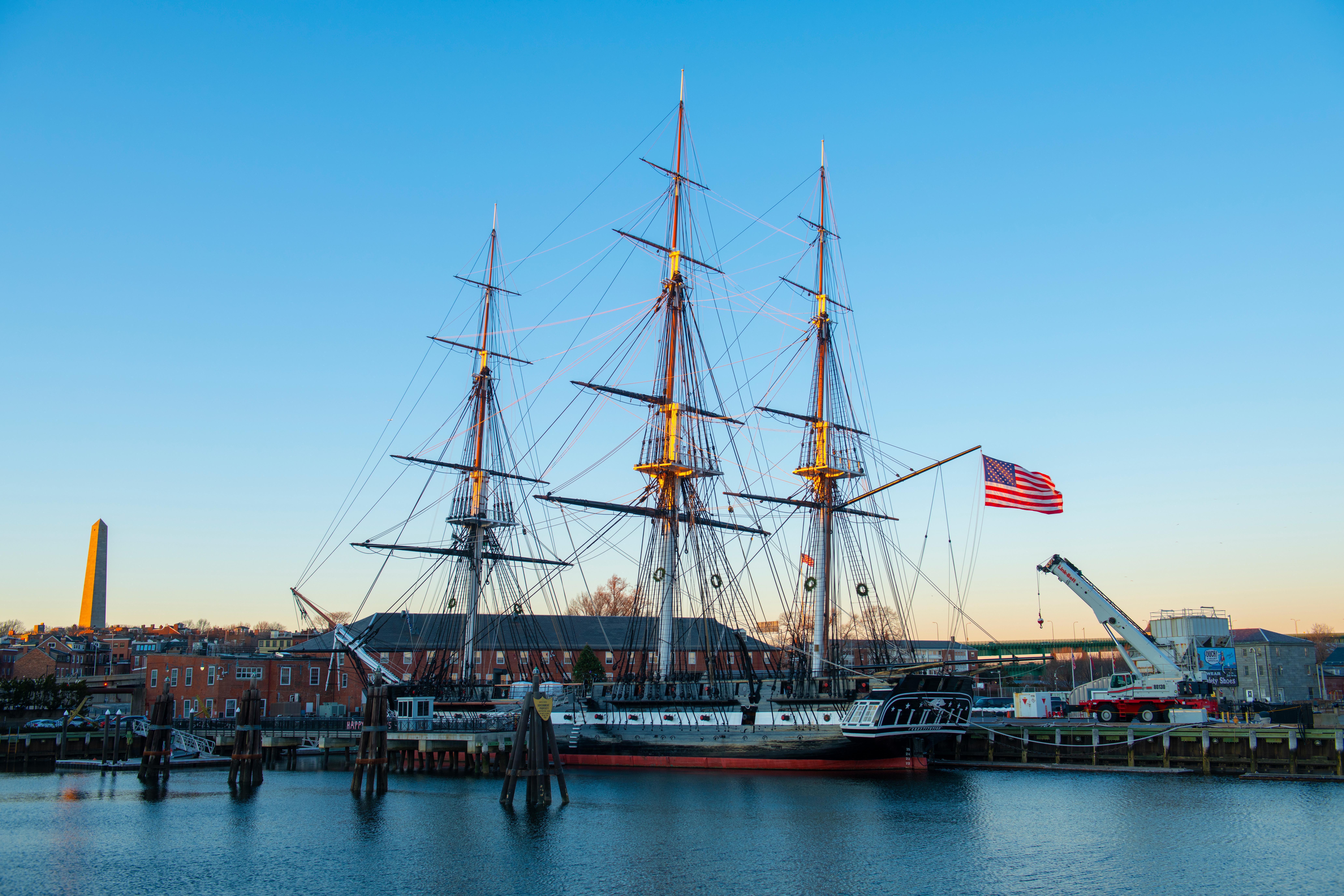 USS Constitution docked in Boston Harbor with American flag and cityscape in the background.