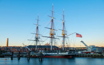 USS Constitution docked in Boston Harbor with American flag and cityscape in the background.