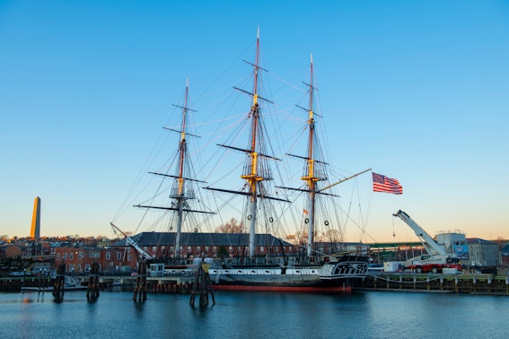 USS Constitution docked in Boston Harbor with American flag and cityscape in the background.