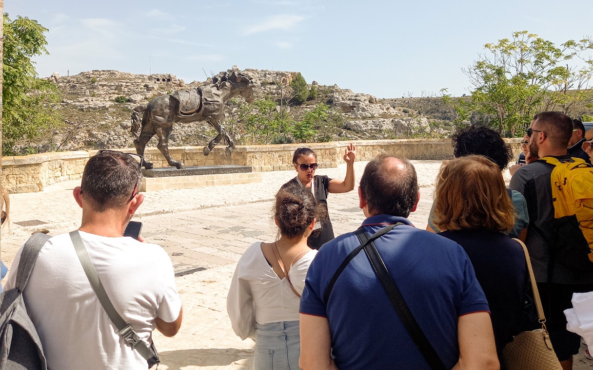 Tour group in Matera listening to a guide near a horse statue, with rocky landscape in the background.