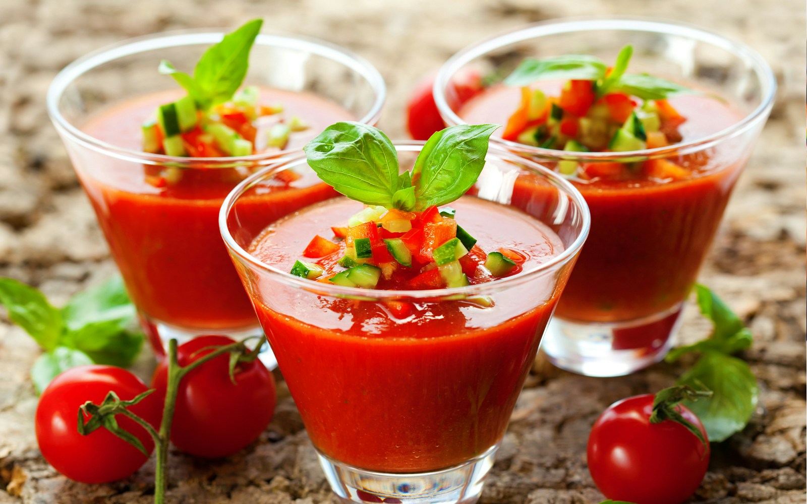 Three glasses of gazpacho garnished with diced vegetables and basil leaves.