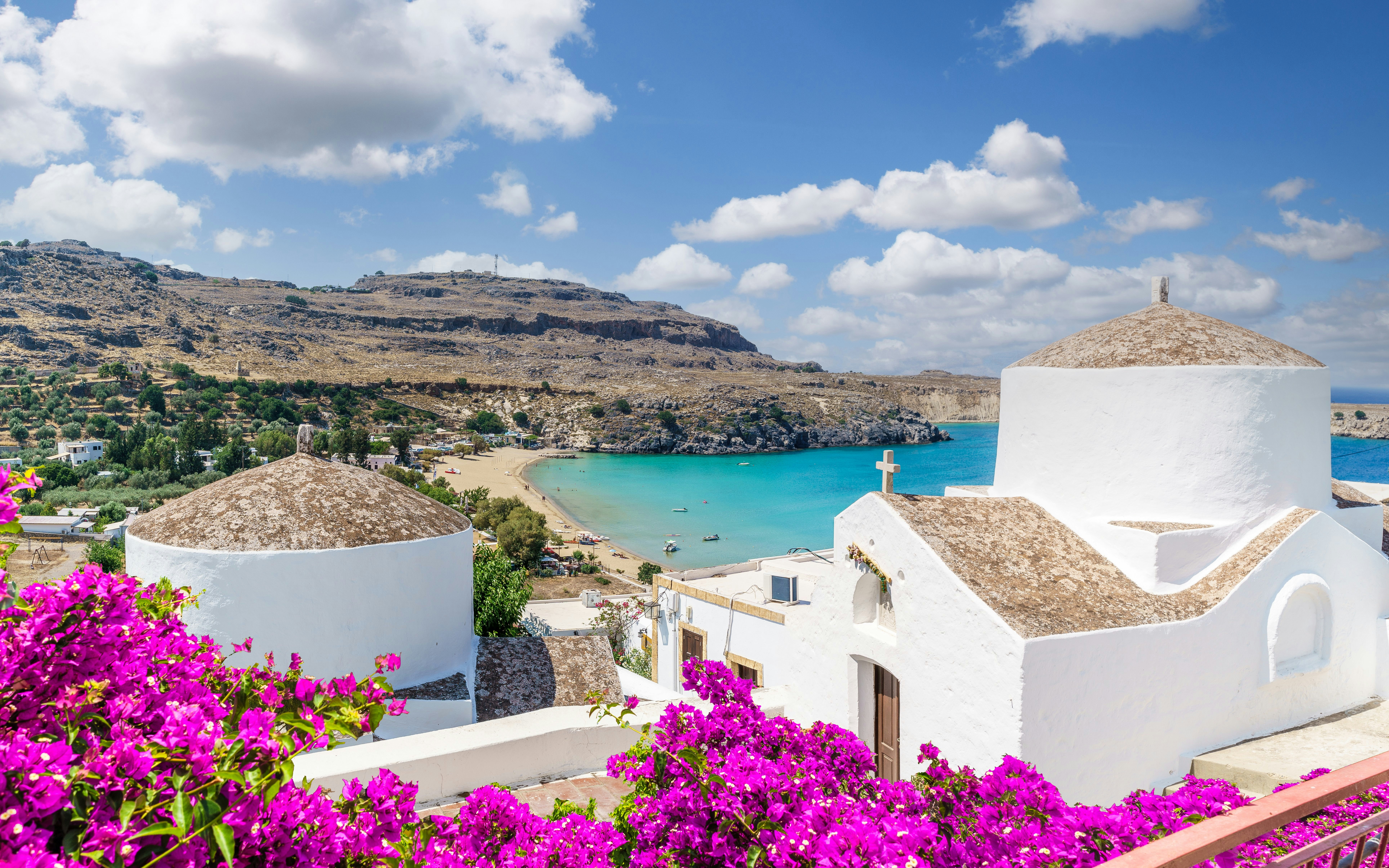 White church with domed roof overlooking Lindos Bay, Rhodes, Greece, surrounded by vibrant bougainvillea.