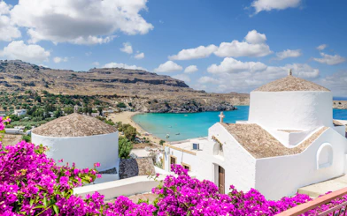 White church with domed roof overlooking Lindos Bay, Rhodes, Greece, surrounded by vibrant bougainvillea.