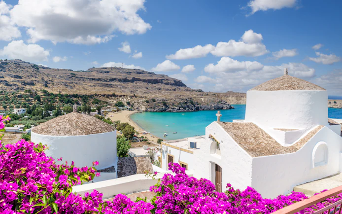 White church with domed roof overlooking Lindos Bay, Rhodes, Greece, surrounded by vibrant bougainvillea.
