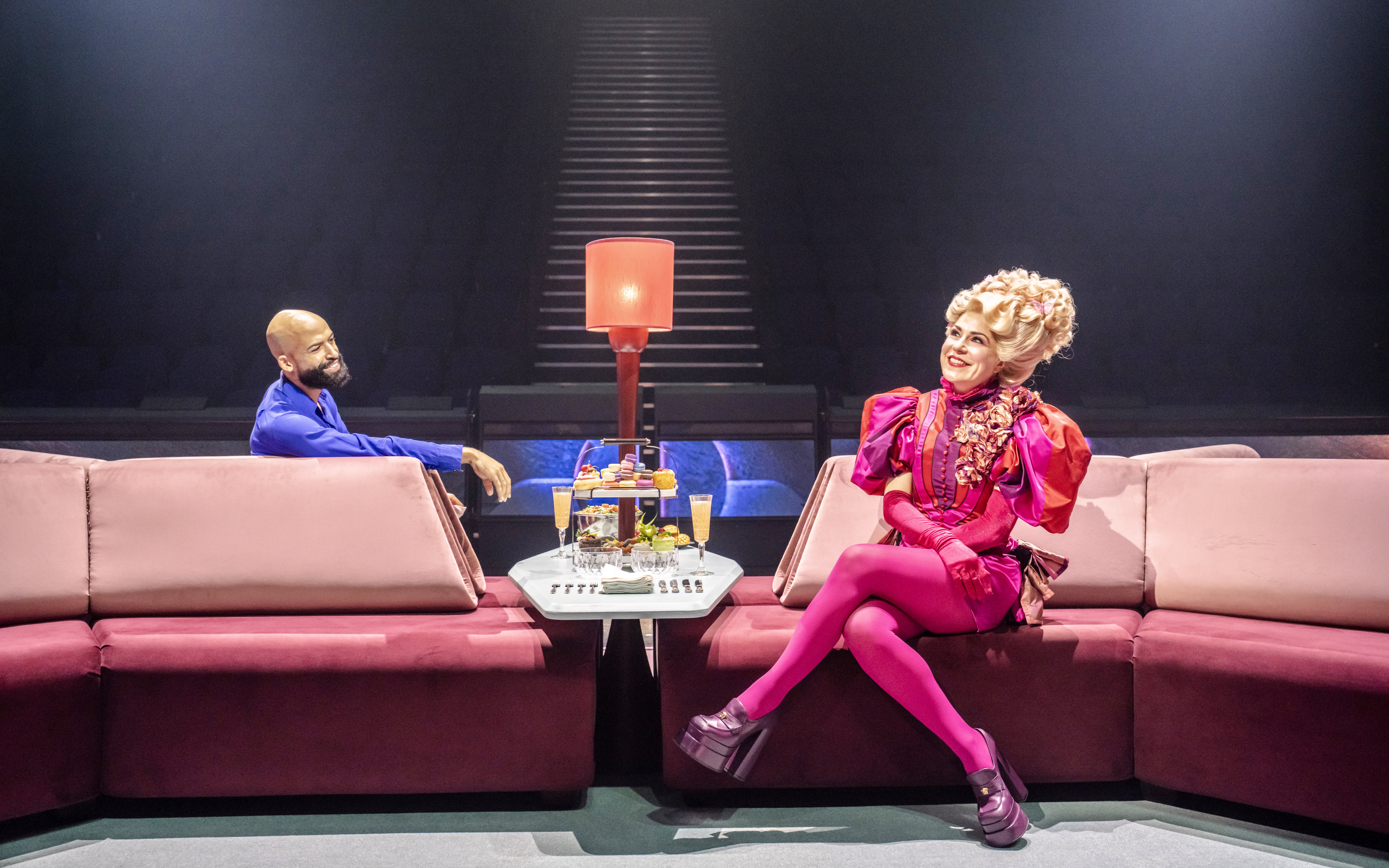 Stage scene from Hunger Games West End show with two actors seated on a pink sofa.