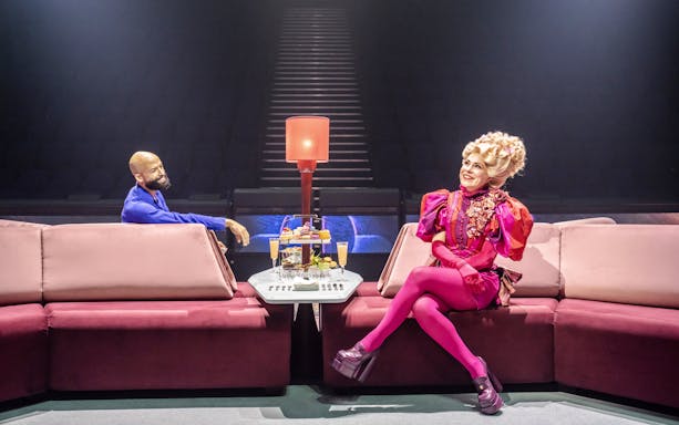 Stage scene from Hunger Games West End show with two actors seated on a pink sofa.