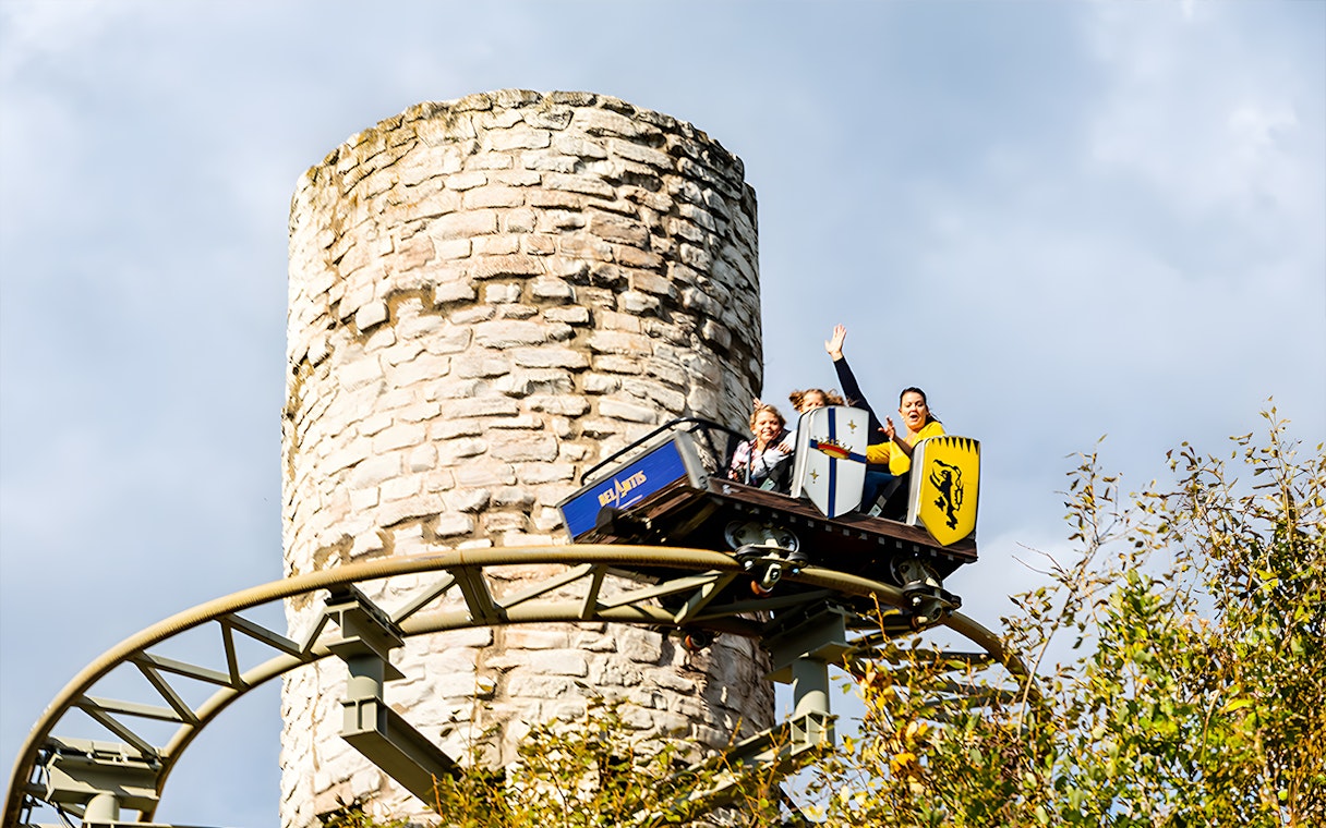 Roller coaster at Belantis Adventure Park with riders near a stone tower.