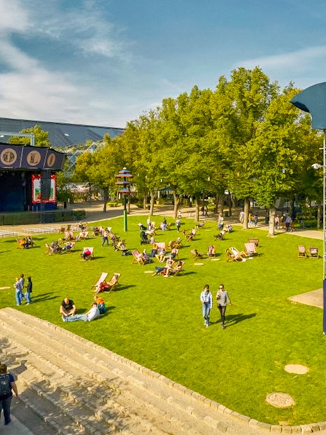 Visitors relaxing on the lawn at Tivoli Gardens, Copenhagen, with nearby attractions and rides.